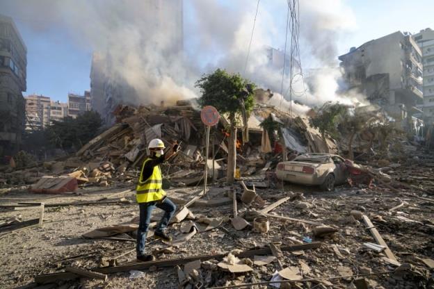 A man in a helmet, covering over his mouth, and reflective vest, gestures while standing on a road with much co<em></em>ncrete debris, and a damaged vehicle.