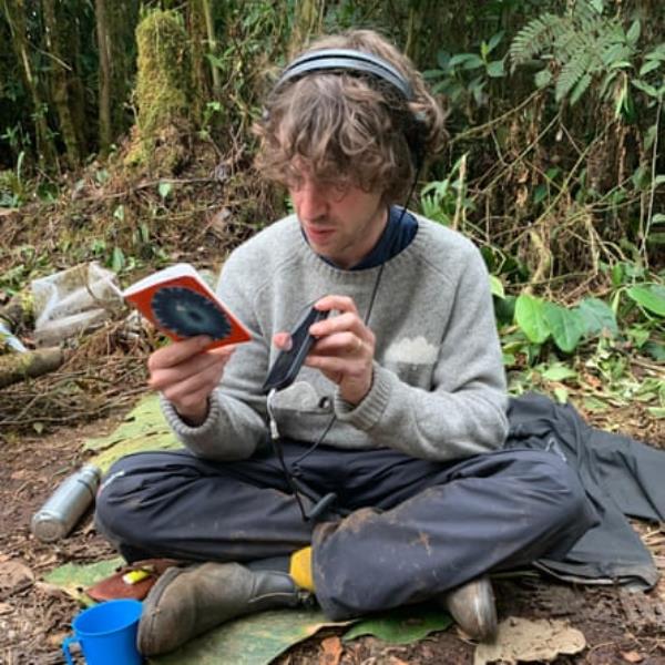 Cosmo Sheldrake is sitting on the forest floor. He is hearing headpho<em></em>nes and holding a recording device