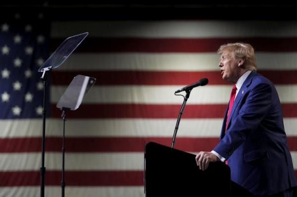 A man is photographed from the side speaking into a microphone with a large American flag as a backdrop.