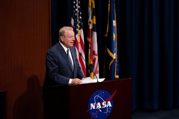 Al Gore in business suit at wooden podium with NASA logo; flags in the background