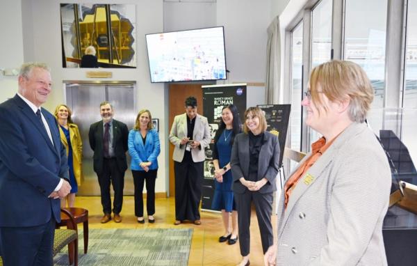 Al Gore in business suit at far left in a brightly lit lobby among a small crowd of NASA Goddard leaders; the floor-to-ceiling glass panes overlooking the high bay clean room is at right